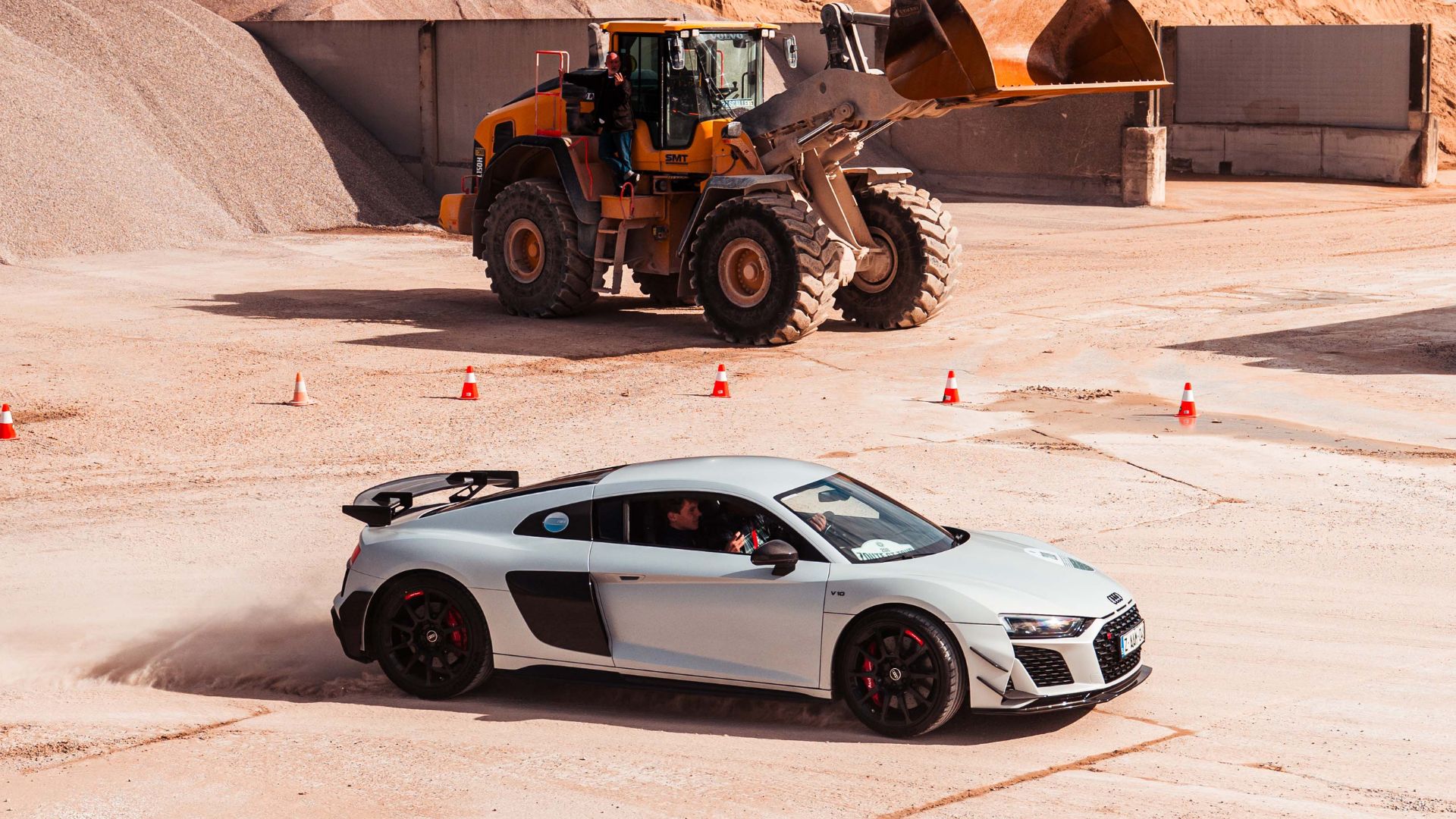Zilverkleurige Audi R8 op een zandpiste voor een wiellader